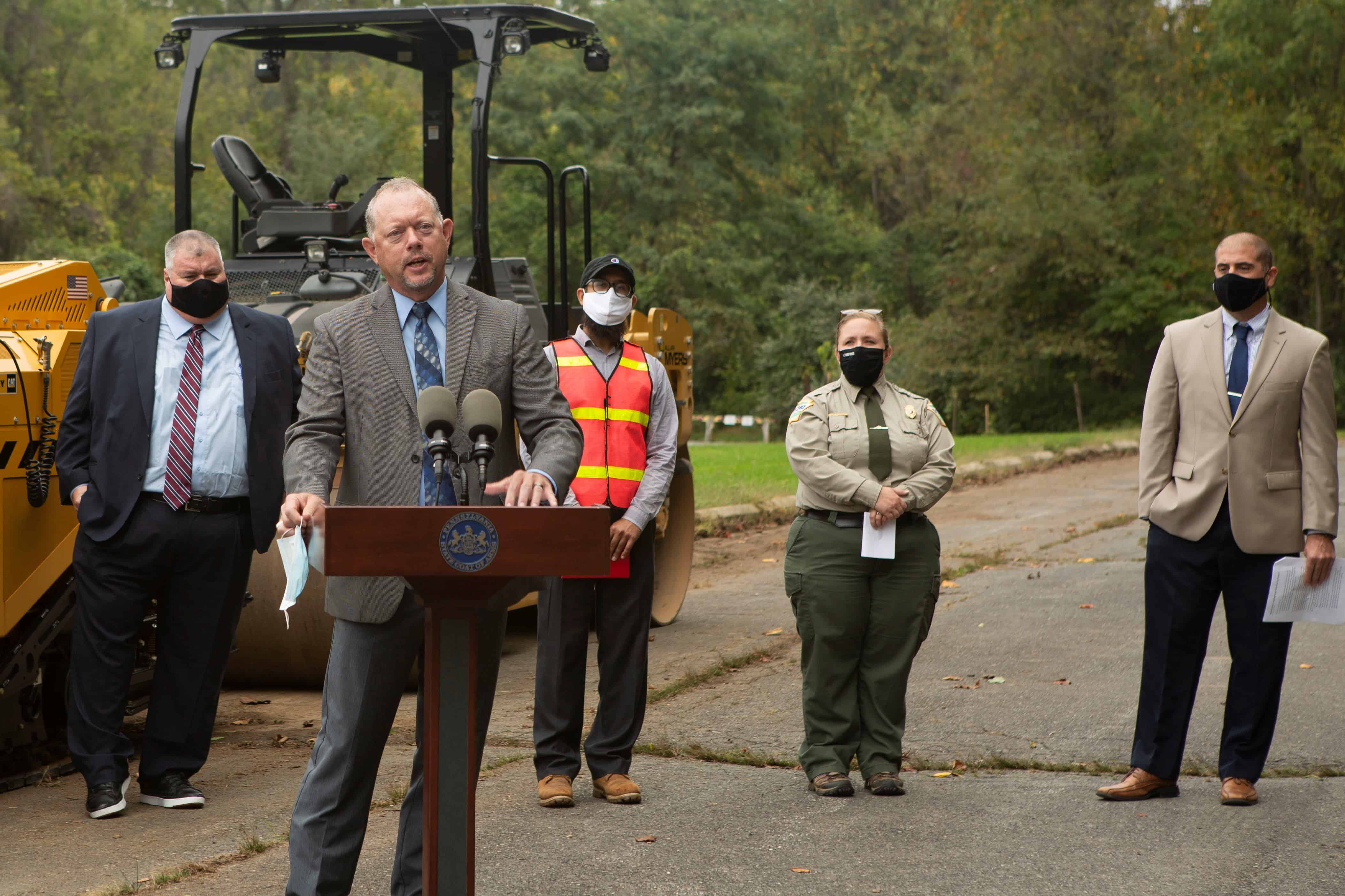 PennDOT's Mike Keiser takes the podium at an event announcing the recycled plastic pilot initiative. Four speakers stand behind him with masks on.